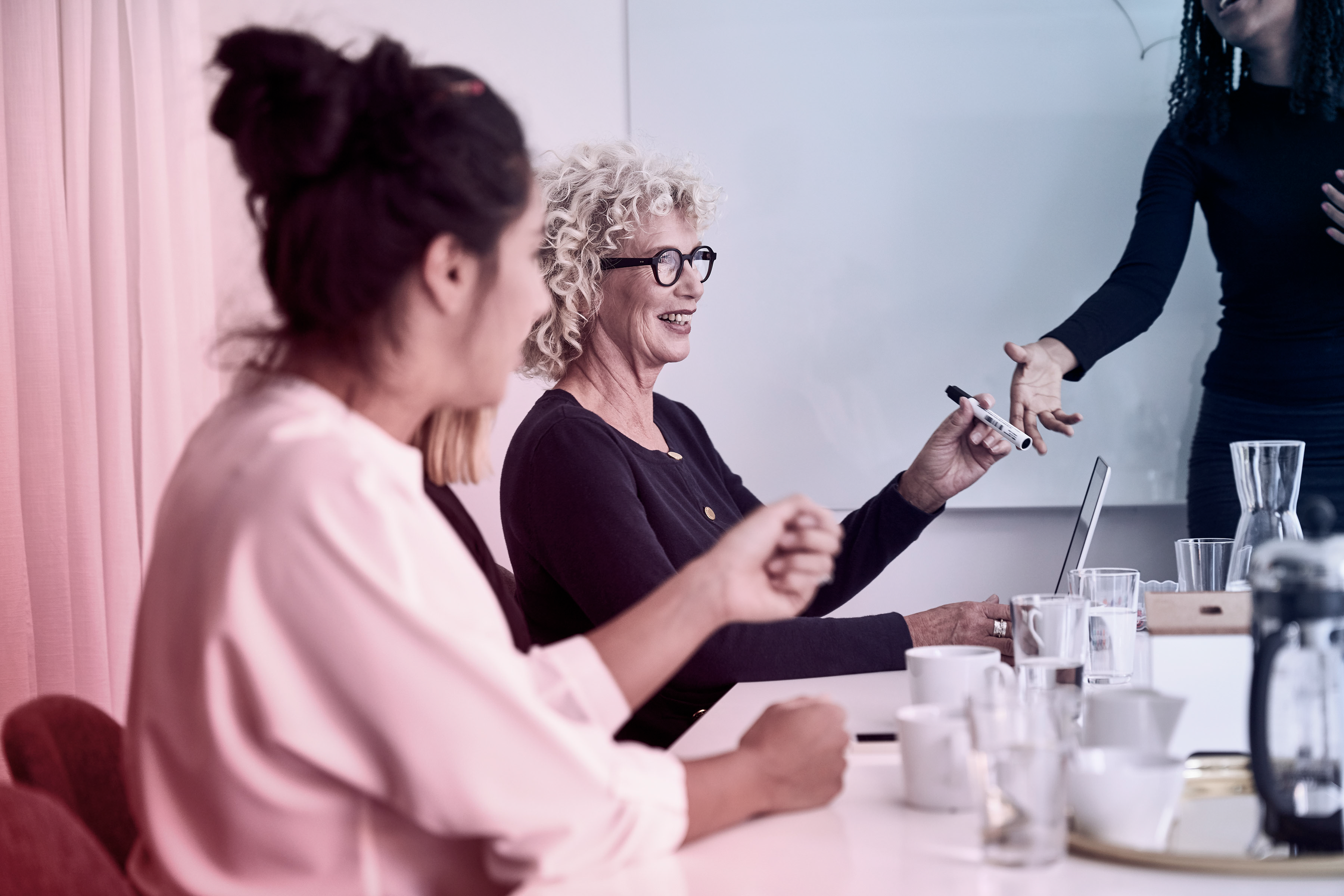 Kvinnor i möte runt ett konferensbord, en av dem ler och antecknar medan en kollega presenterar vid whiteboard.