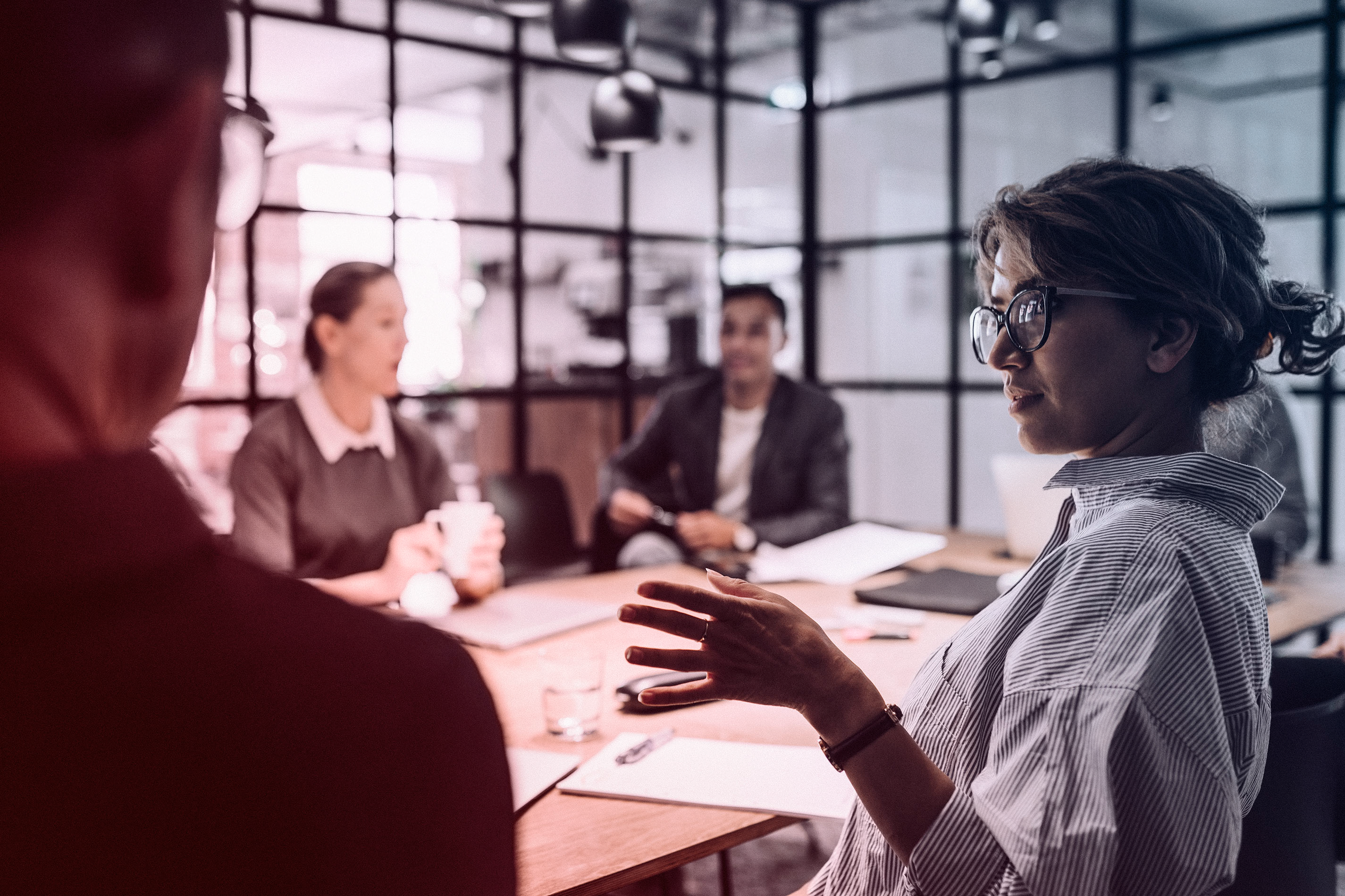 A woman in glasses speaks during a meeting in a modern office, while three colleagues listen attentively around the conference table.