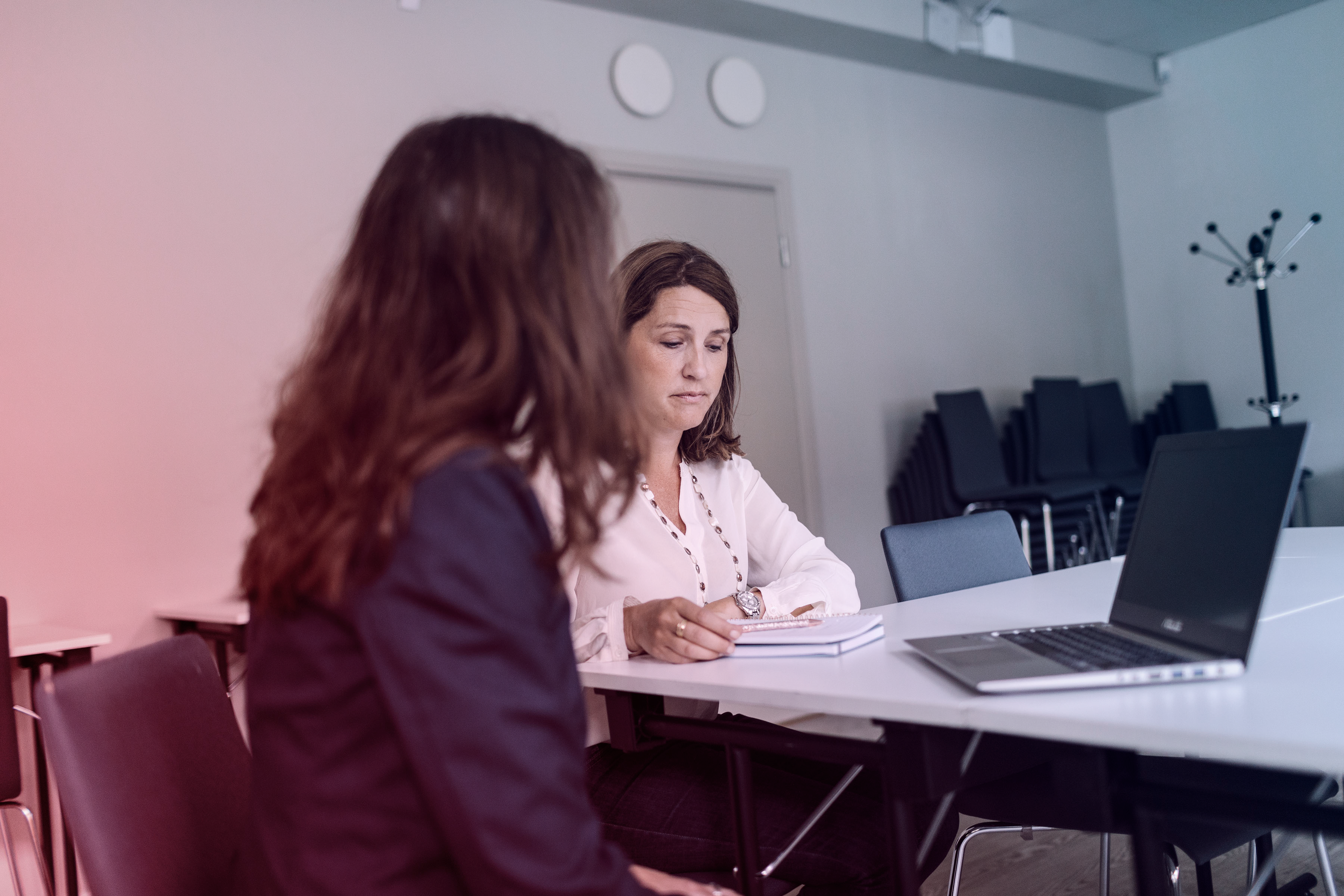 Two women in a meeting room, sitting at a table with a laptop and notebook, engaged in a serious discussion.