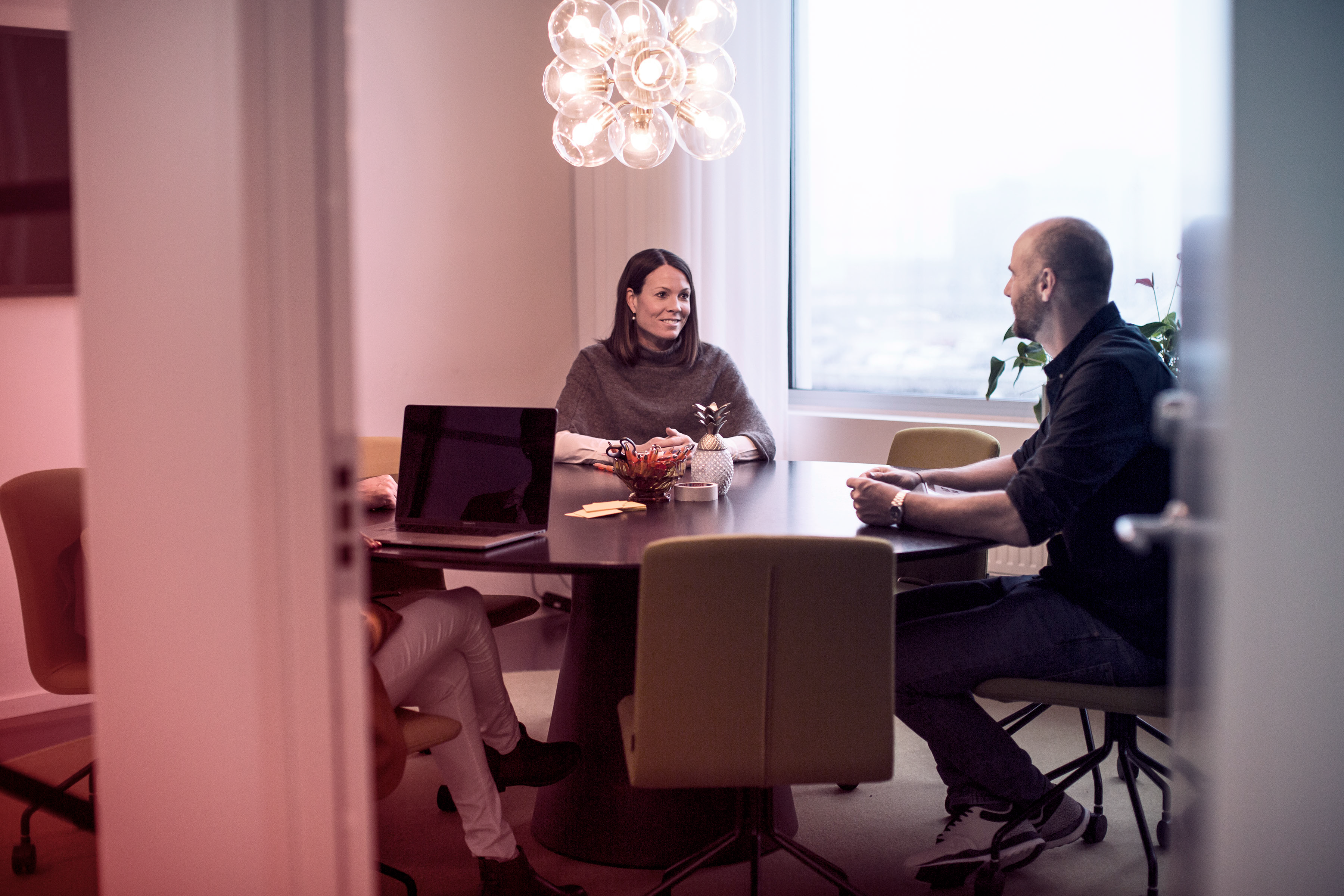 Tre personer i ett modernt mötesrum samtalar runt ett runt bord, med laptop, anteckningar och snacks framför sig – en avslappnad men professionell stämning som speglar arbetsmiljö och teamdynamik.